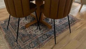 Une table de salle à manger ronde en bois, entourée de six chaises, un vase contenant des fleurs séchées et un tapis à motifs posé sur un plancher en bois.