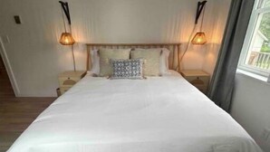 Un grand lit recouvert d’une couvre-lit blanche, deux lampes de chevet, un banc en bois et un tapis à motifs.
