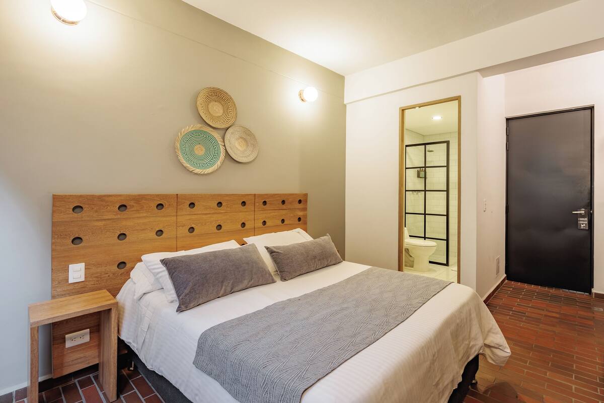 Cama bem arrumada com cabeceira de madeira, mesa de cabeceira e parede com pratos decorativos.