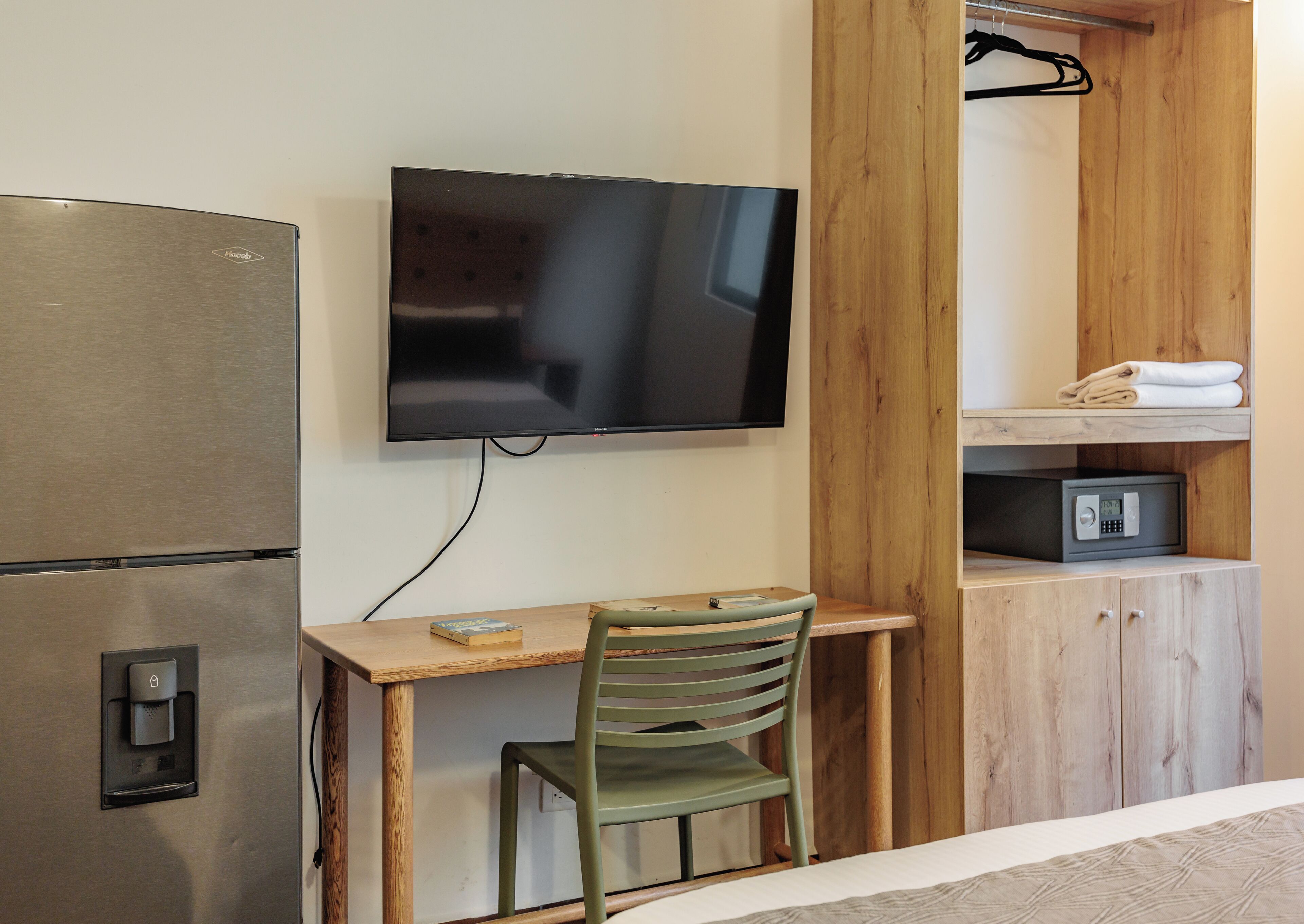A compact hotel room with a kitchenette, a flat-screen TV mounted on the wall, a wooden desk with a chair, and a small safe on a wooden cabinet.