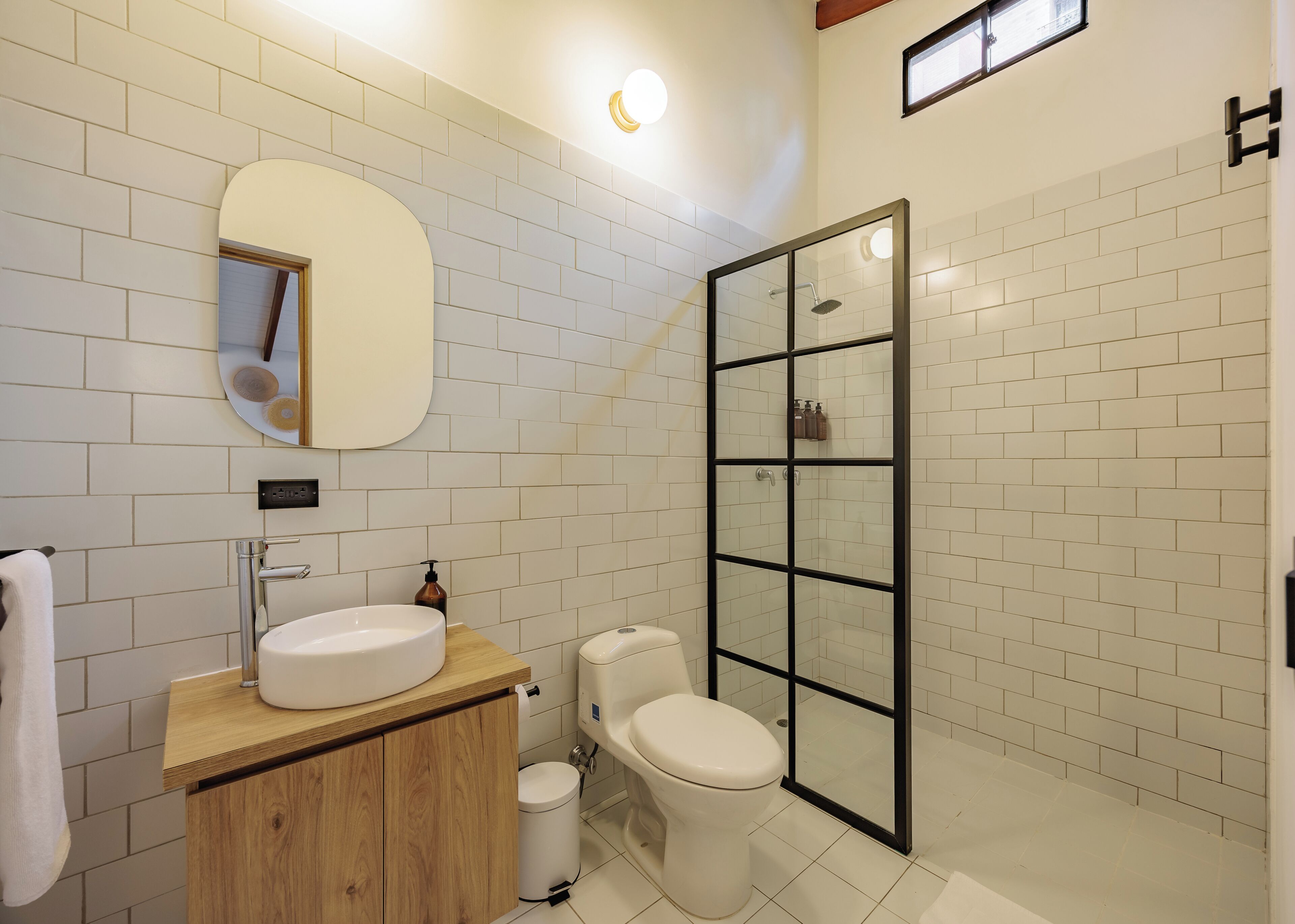 Un bagno con parete rivestita in piastrelle bianche, un mobile vanitario in legno, uno specchio rotondo, un water e una cabina doccia con pareti in vetro.