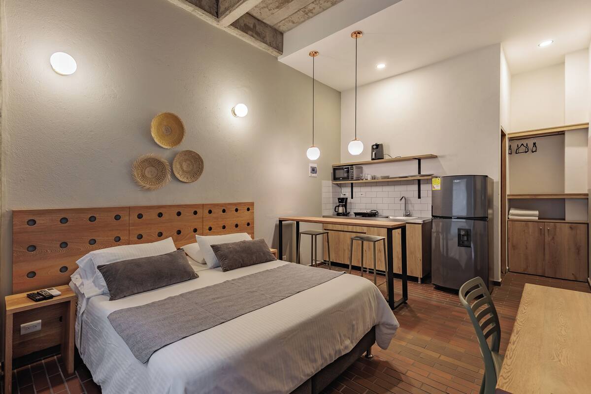 Quarto de hotel pequeno com cama, mesa de cabeceira e uma kitchenette.