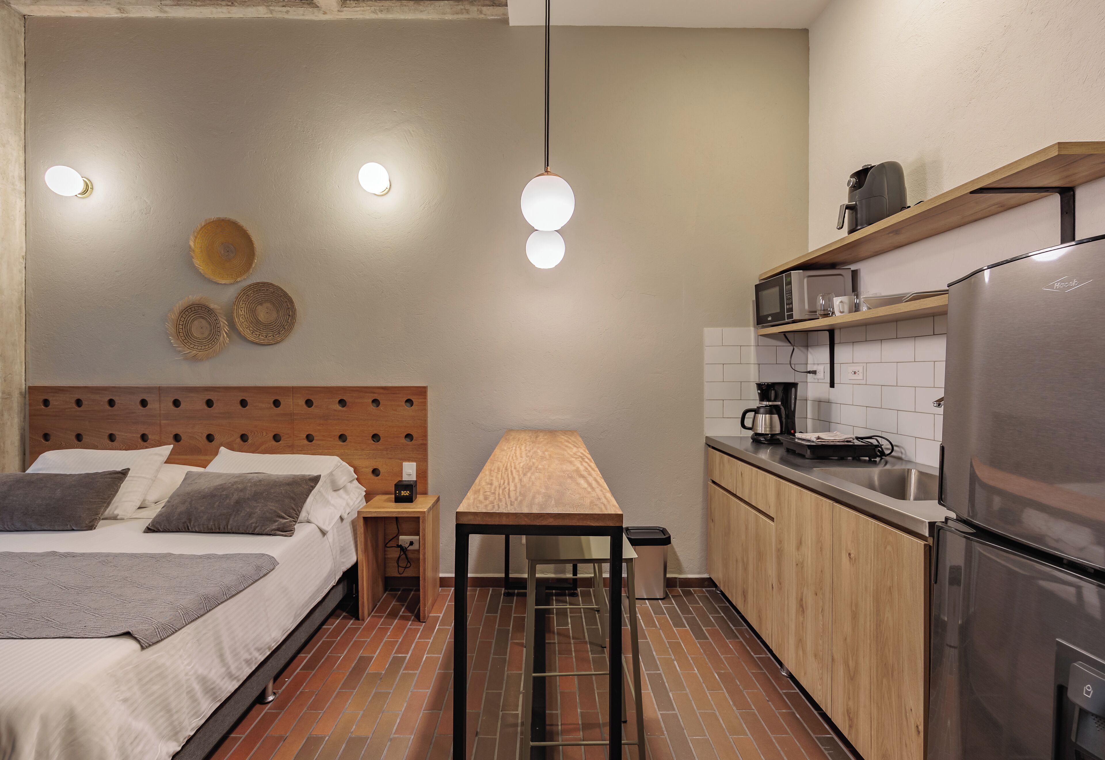 Un dormitorio compacto con cama, mesita de noche y una pequeña mesa de madera. Se ve una kitchenette con refrigerador, microondas y cafetera.