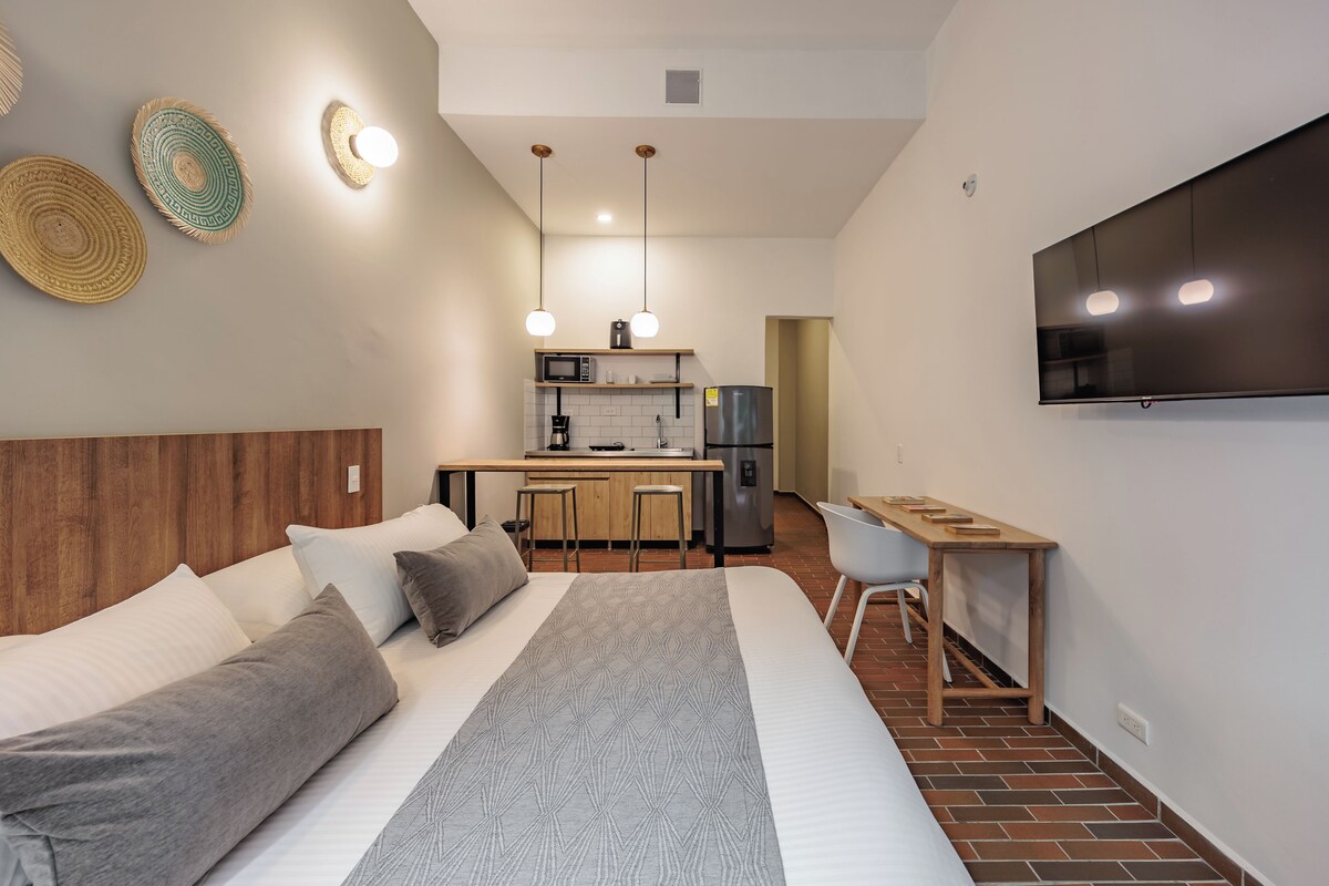 A modern hotel room with a large bed, a desk, a kitchenette, and a TV mounted on the wall.