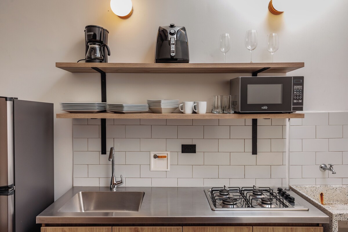 Uma cozinha moderna com pia de aço inoxidável, fogão a gás, micro-ondas e prateleiras de madeira com cafeteira, pratos e copos.