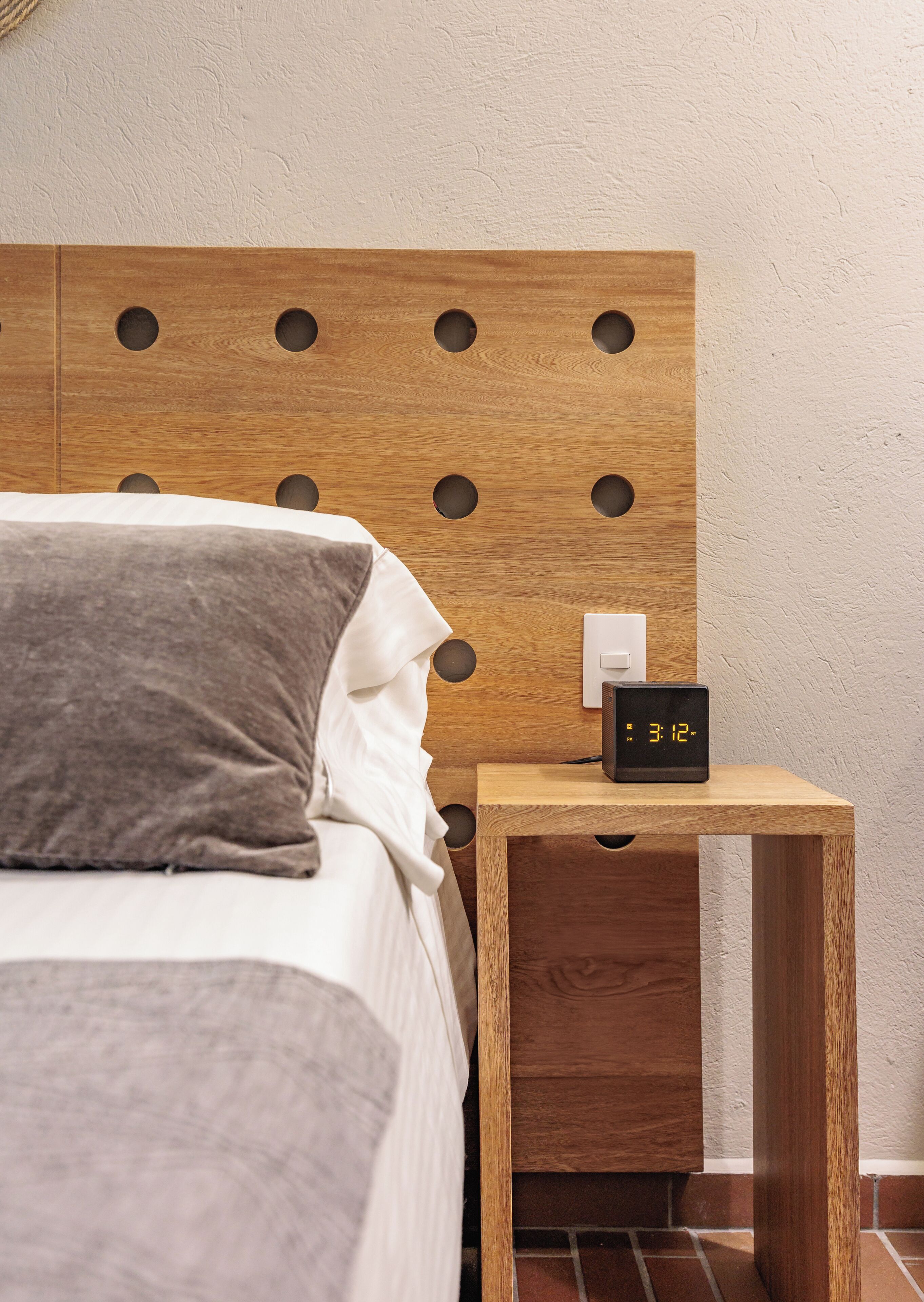 Un lit bien fait, doté d’une tête de lit en bois ornée de découpes circulaires, une table de chevet avec une horloge numérique et un mur blanc en arrière-plan.