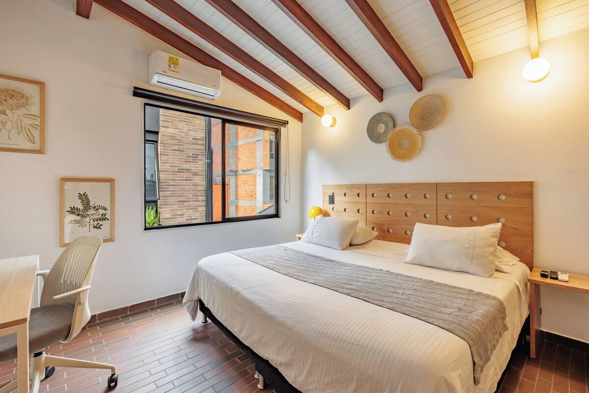 Quarto com cama grande, escrivaninha e cadeira. Há uma janela com persianas, ar-condicionado fixado na parede e teto com vigas aparentes.