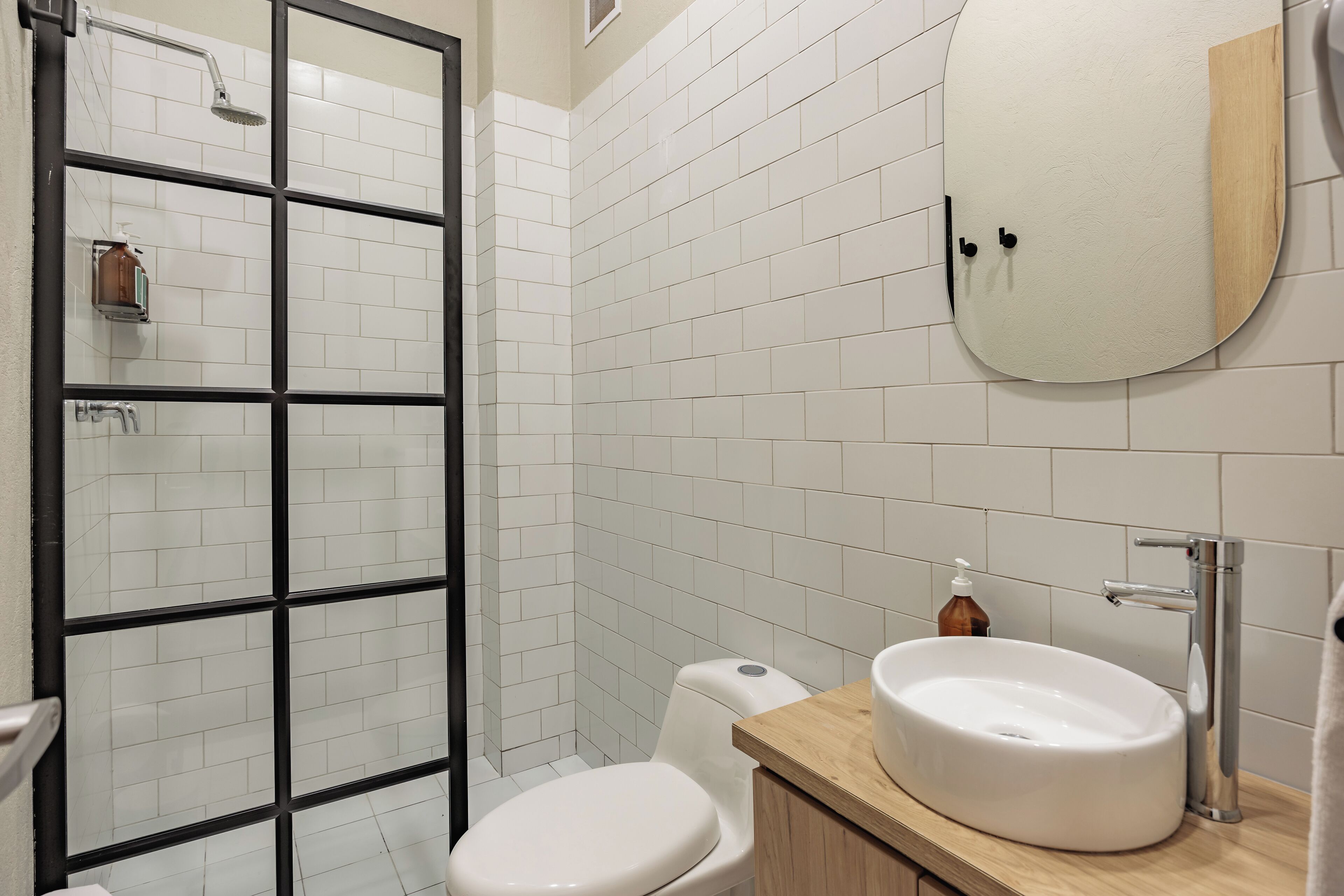 A bathroom with white tiled walls, a wooden vanity, a white sink, a toilet, and a shower with a glass door.
