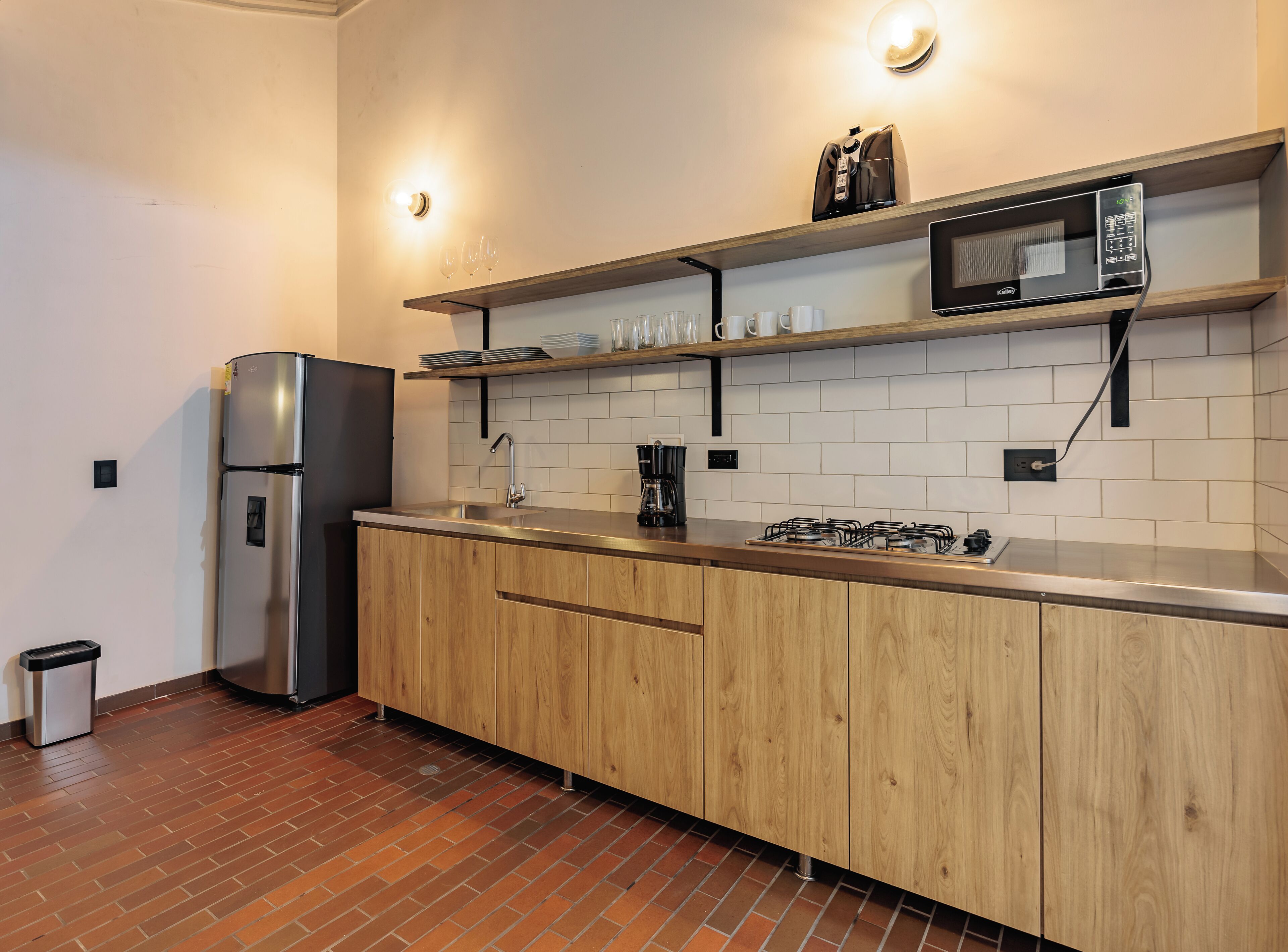 A modern kitchen with a stainless steel refrigerator, a microwave, and a gas stove. There are wooden cabinets and a brick floor.