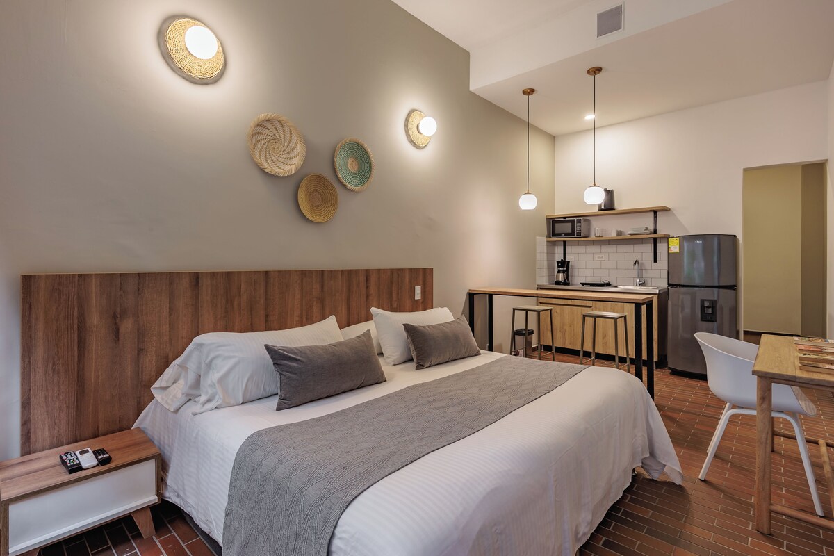 Quarto de hotel moderno com cabeceira de madeira, cama grande com roupa de cama branca e cinza, mesa de cabeceira pequena com telefone, cozinha compacta com micro-ondas, frigorífico e banquetas.