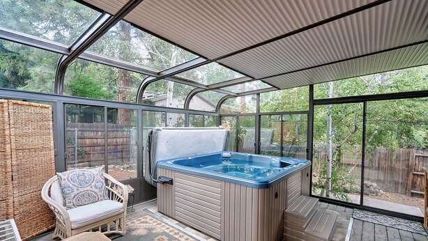 A glass-enclosed patio with a hot tub, wicker furniture, and a view of a wooded area. - Forest Heights (Flagstaff)