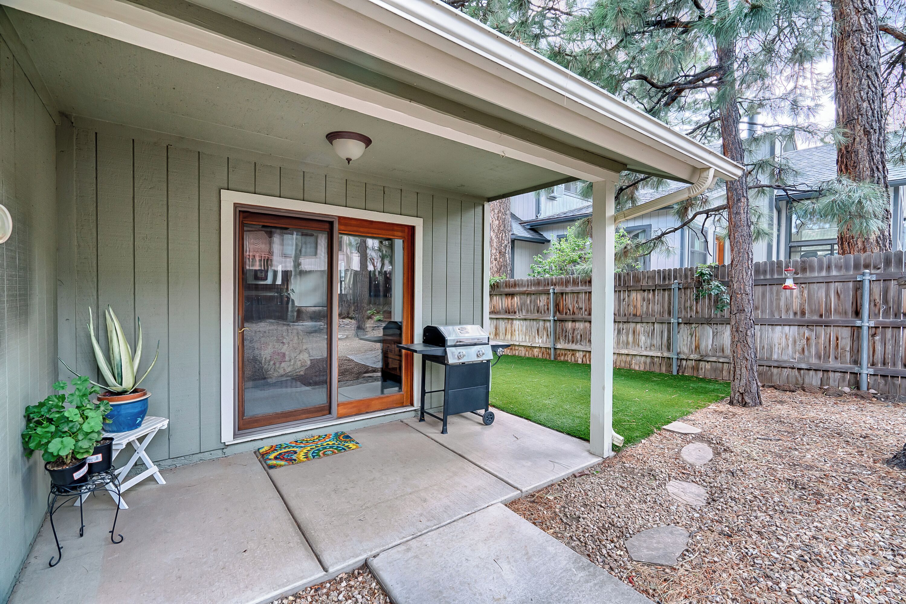 A covered patio with a grill, potted plants, and a wooden fence in the background.