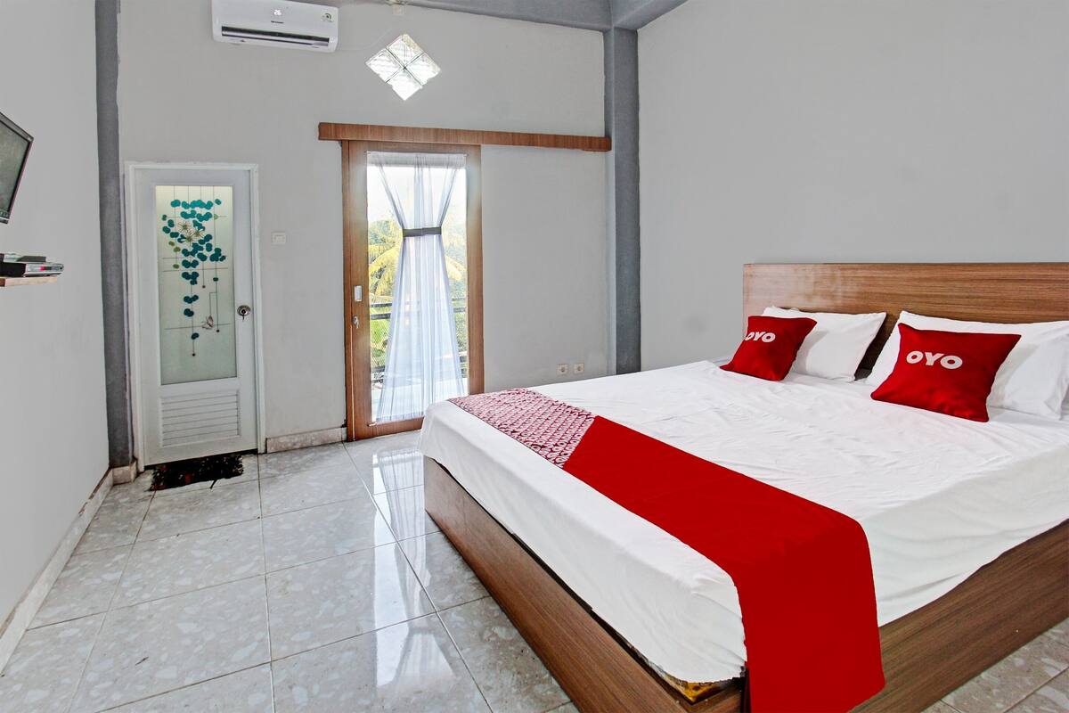 Une chambre d’hôtel avec un lit, une porte dotée d’un panneau en verre dépoli, une fenêtre avec des rideaux et un sol carrelé.