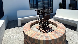A circular brick firepit with a metal grill in front of a white building with large windows and a sign that reads "Villa Fantastica 11 Kalkoond Crescent."