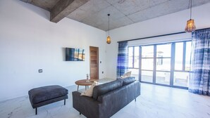 A spacious living area with a grey sofa, a small ottoman, and a round table. There is a large window with blue curtains, a wooden door, and a hanging light fixture.