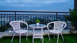 A balcony with two white wicker chairs and a small round table, a potted plant, and a view of a cityscape at dusk. - Luxurious 2 Bed Garden City (Nairobi)