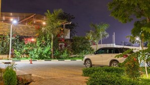 A well-lit parking area with a white car parked, surrounded by lush greenery and a building with a modern design. - Luxurious 2 Bed Garden City (Nairobi)
