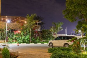 A well-lit parking area with a white car parked, surrounded by lush greenery and a building with a modern design. - Luxurious 2 Bed Garden City (Nairobi)