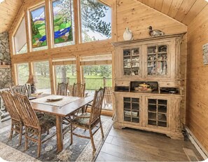A wooden dining area with a table and chairs, a cabinet with glass doors, and large windows with a view of greenery.
