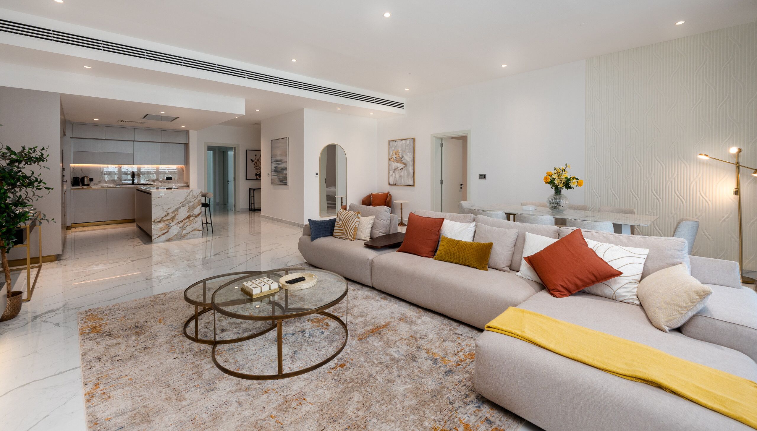 A modern living room with a marble floor, a glass coffee table, and a beige sofa with orange cushions.