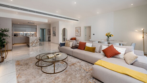 Een moderne woonkamer met een marmeren vloer, een glazen salontafel en een beige bank met oranje kussens.
