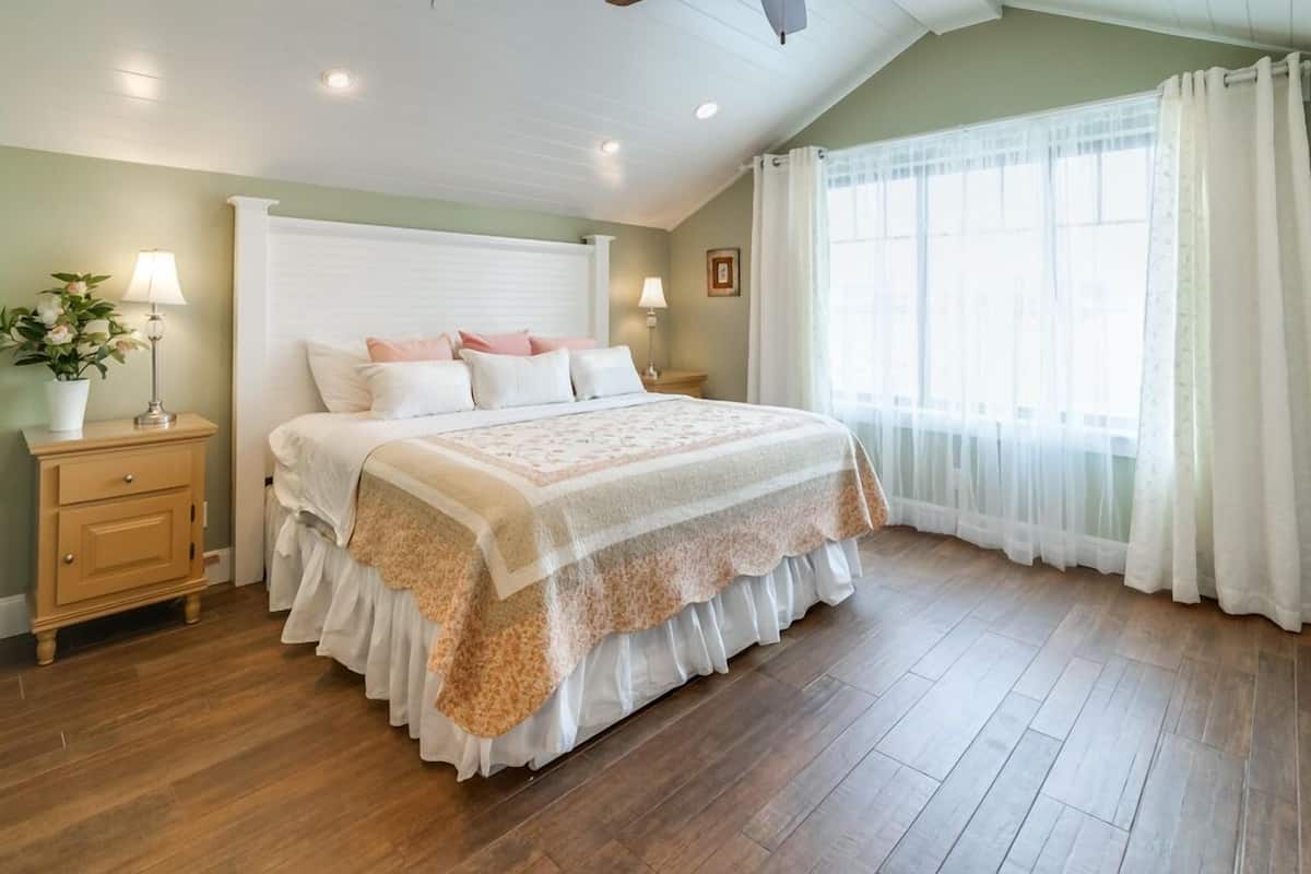 A bedroom with a large bed, wooden flooring, two bedside tables with lamps, a window with sheer curtains, and a wall-mounted light fixture.