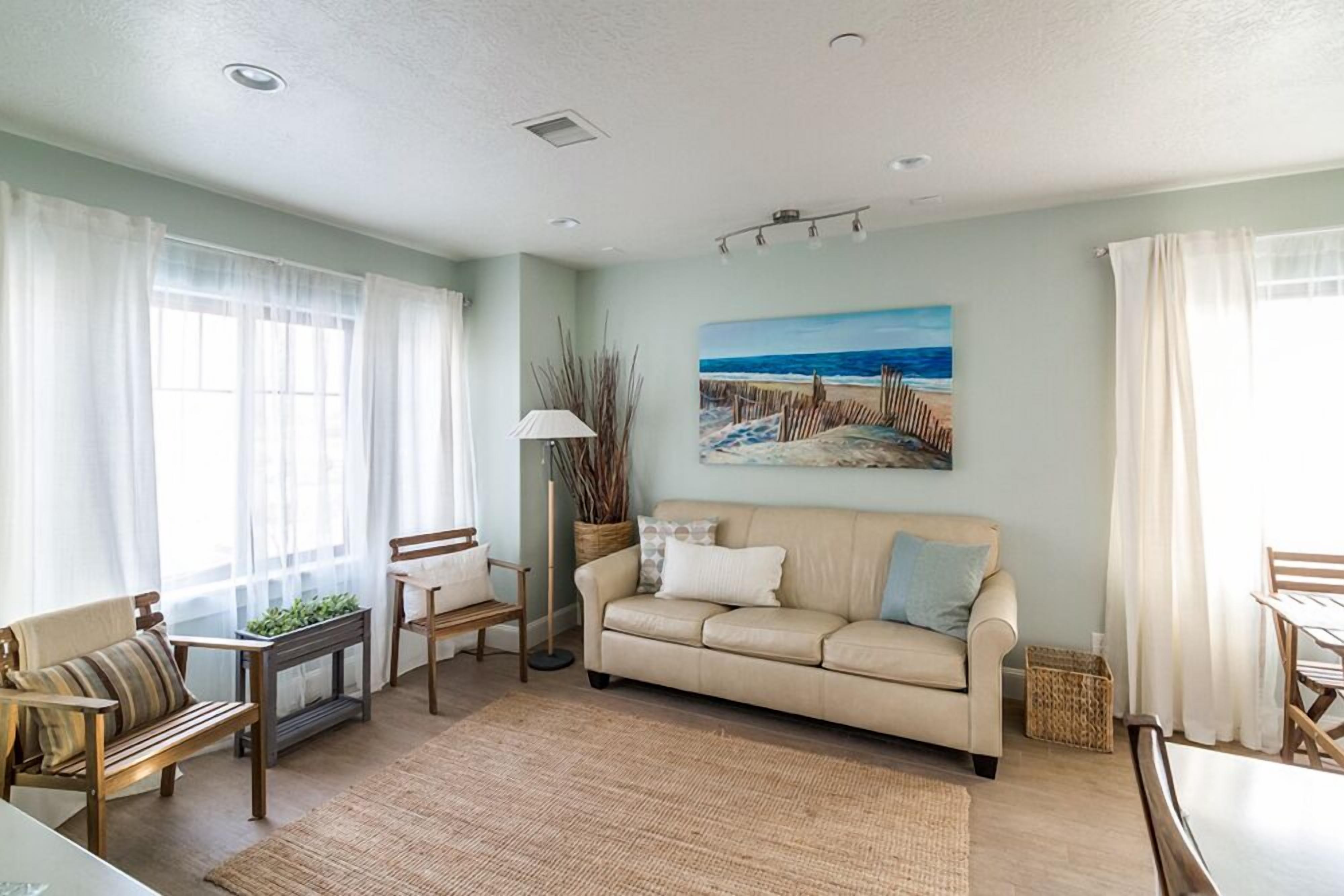 Sala de estar com sofá bege, cadeiras de madeira, uma mesinha e um quadro com uma paisagem de praia.