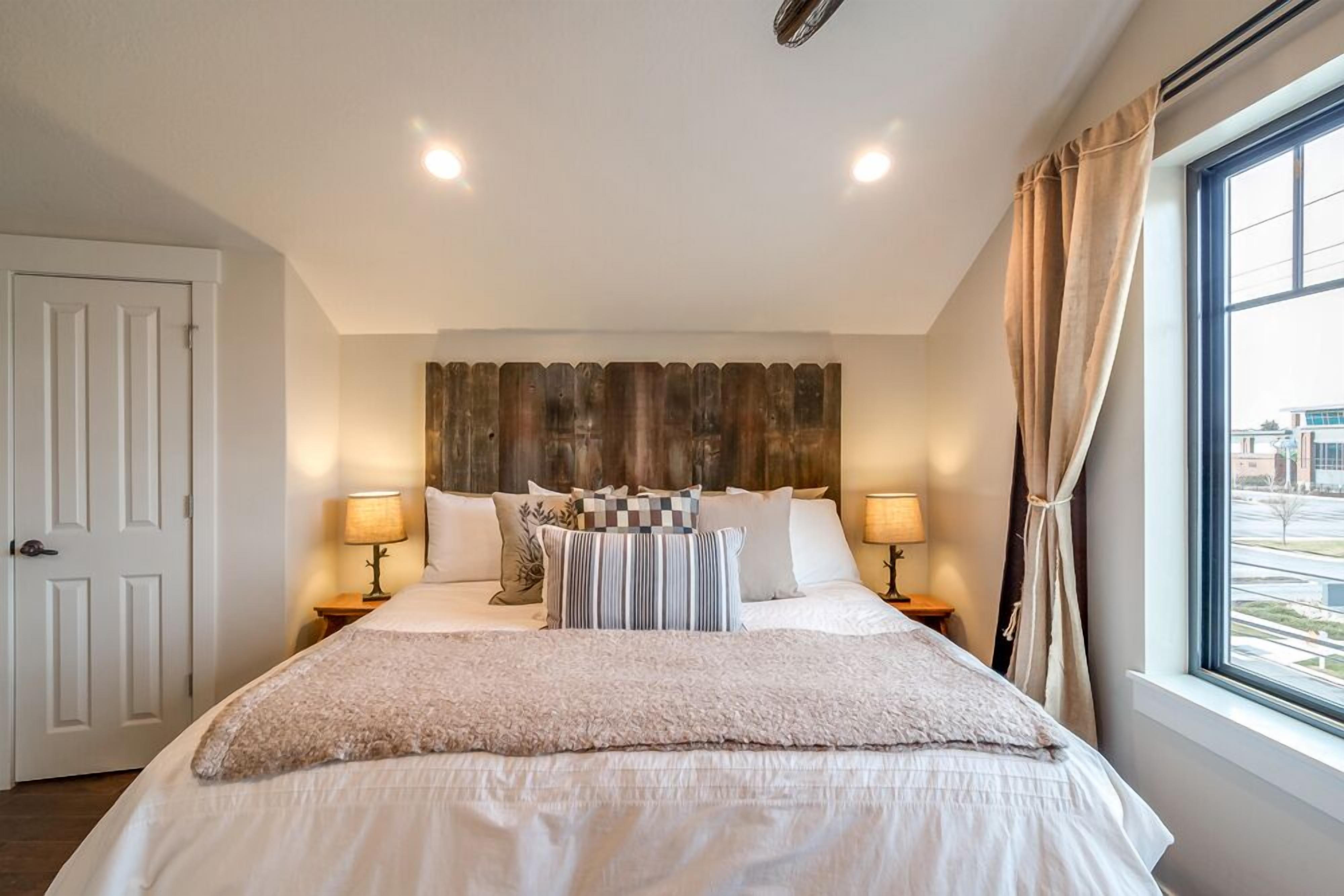 A bedroom with a large bed, two bedside tables with lamps, a wooden headboard, and a window with curtains.