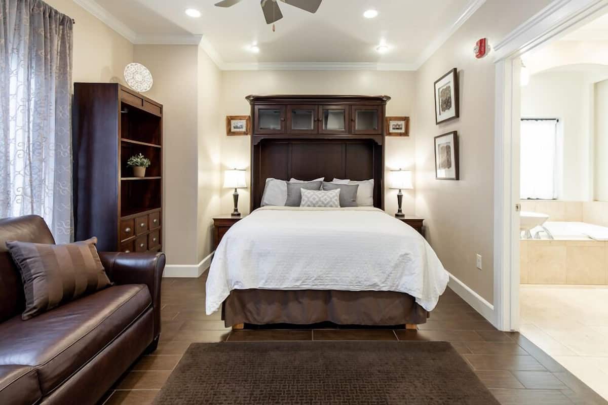 A bedroom with a large bed, a brown leather sofa, a wooden cabinet, and a bathroom visible through an open door.