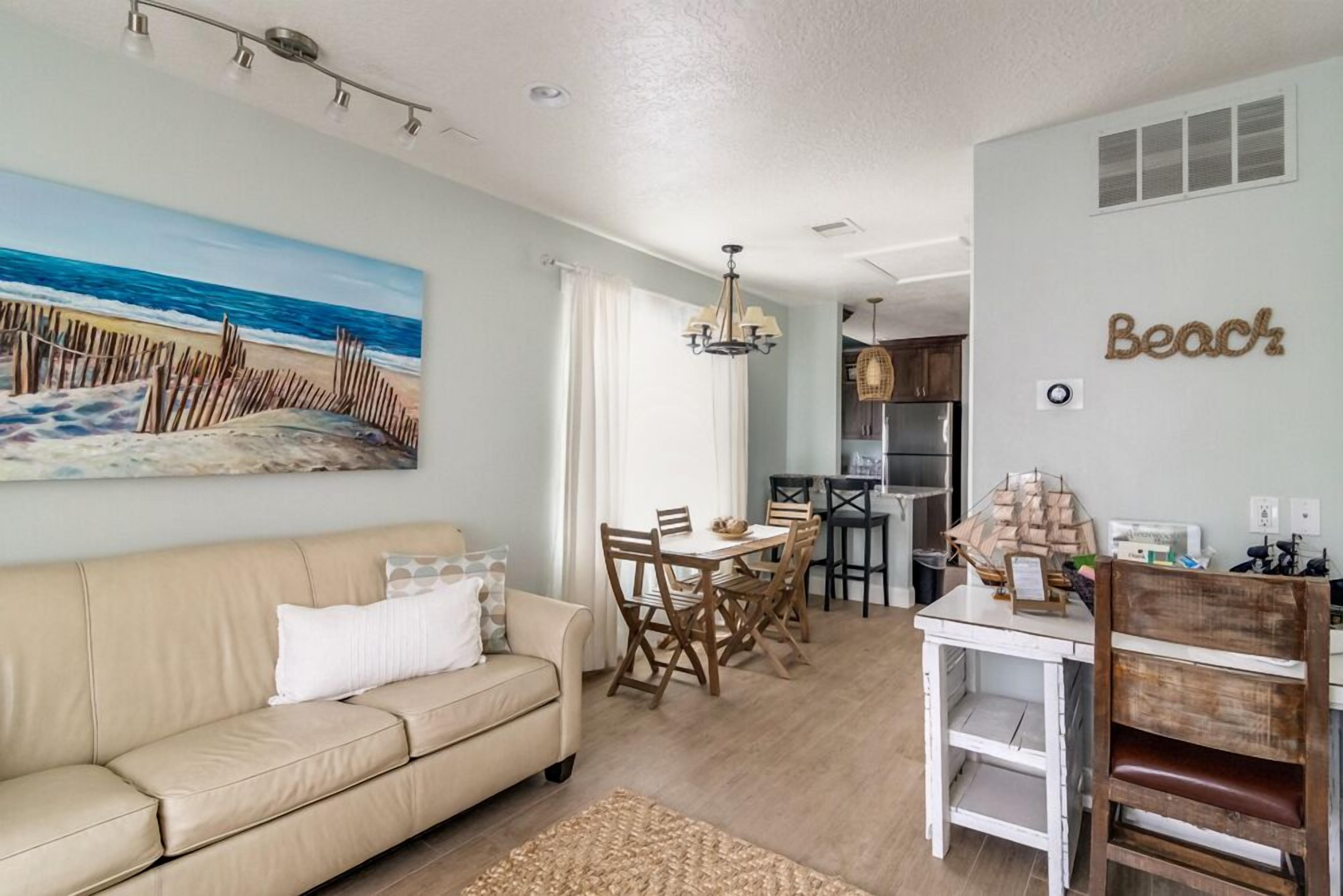 A living room with a beige sofa, a dining table with chairs, a kitchen area, and a beach-themed wall decoration.