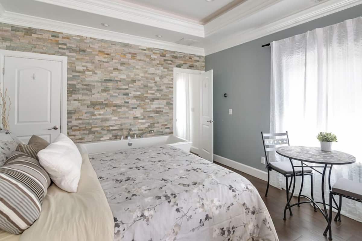A bedroom with a large bed, a stone wall, a window with curtains, and a small round table with chairs.