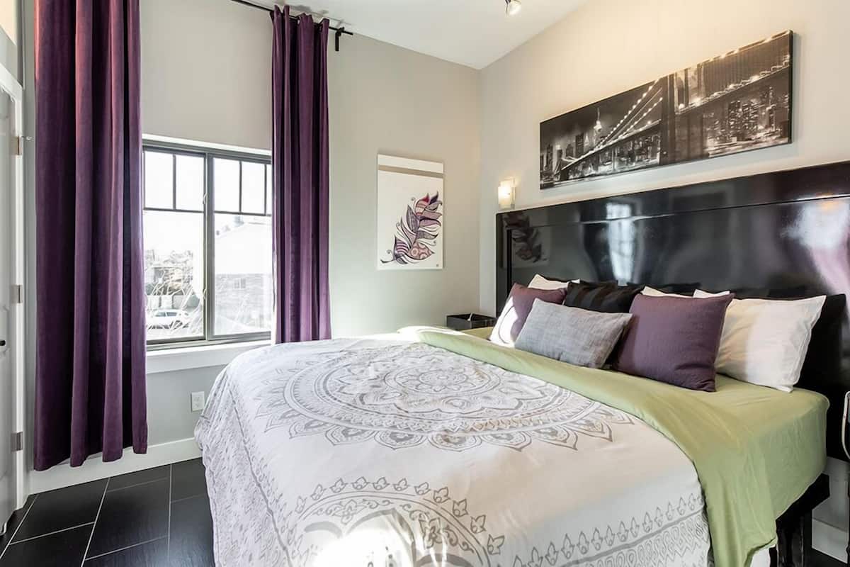 A modern bedroom with a large bed, purple curtains, a cityscape artwork, and a headboard with multiple pillows.