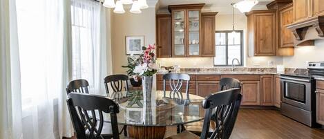 A dining area with a round glass table surrounded by chairs, wooden cabinets, and a kitchen in the background.