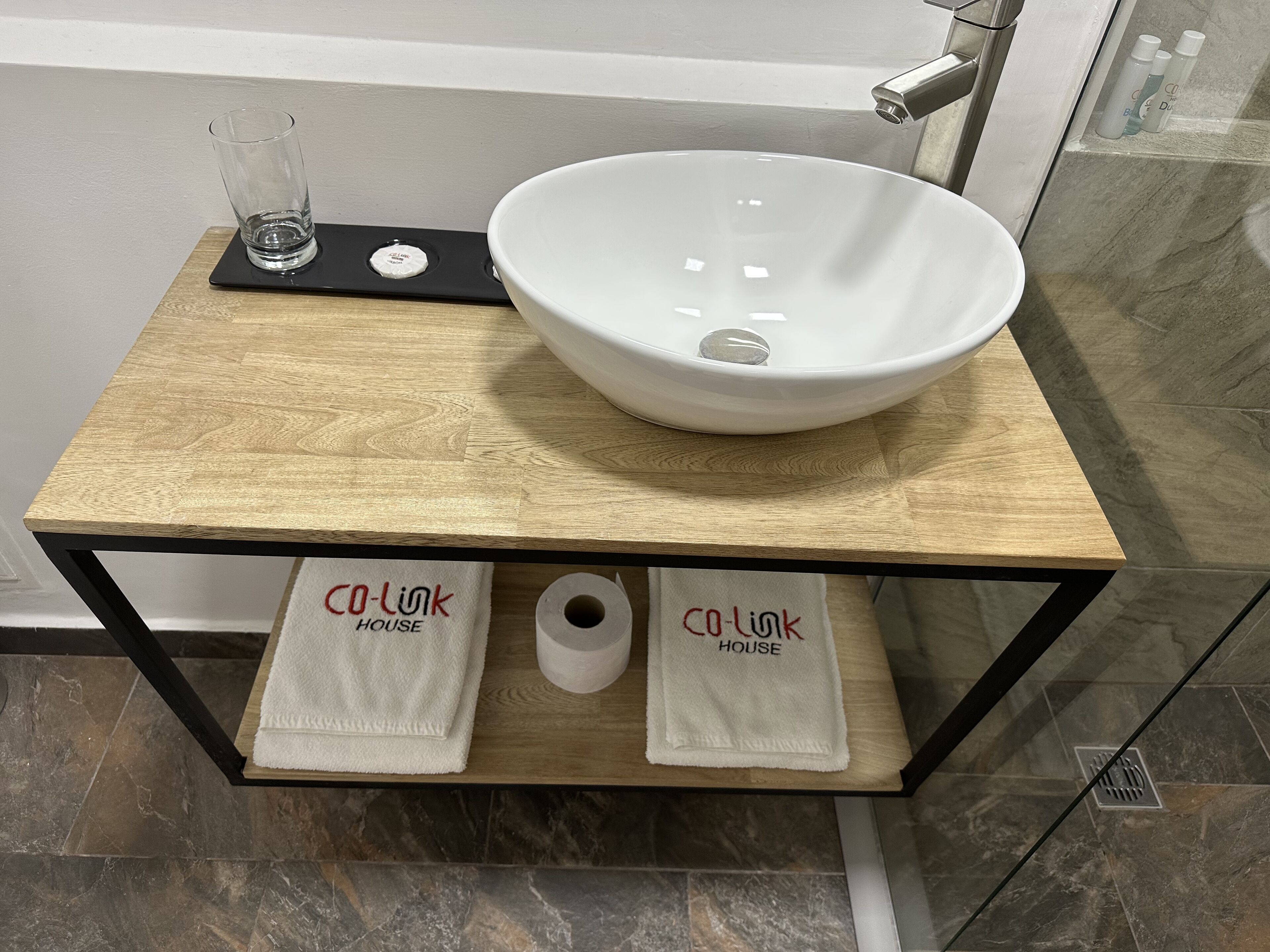 Ein Badezimmer mit einem Holzbadezimmerhocker, einem weißen Waschbecken, zwei Handtüchern und einem Glas.