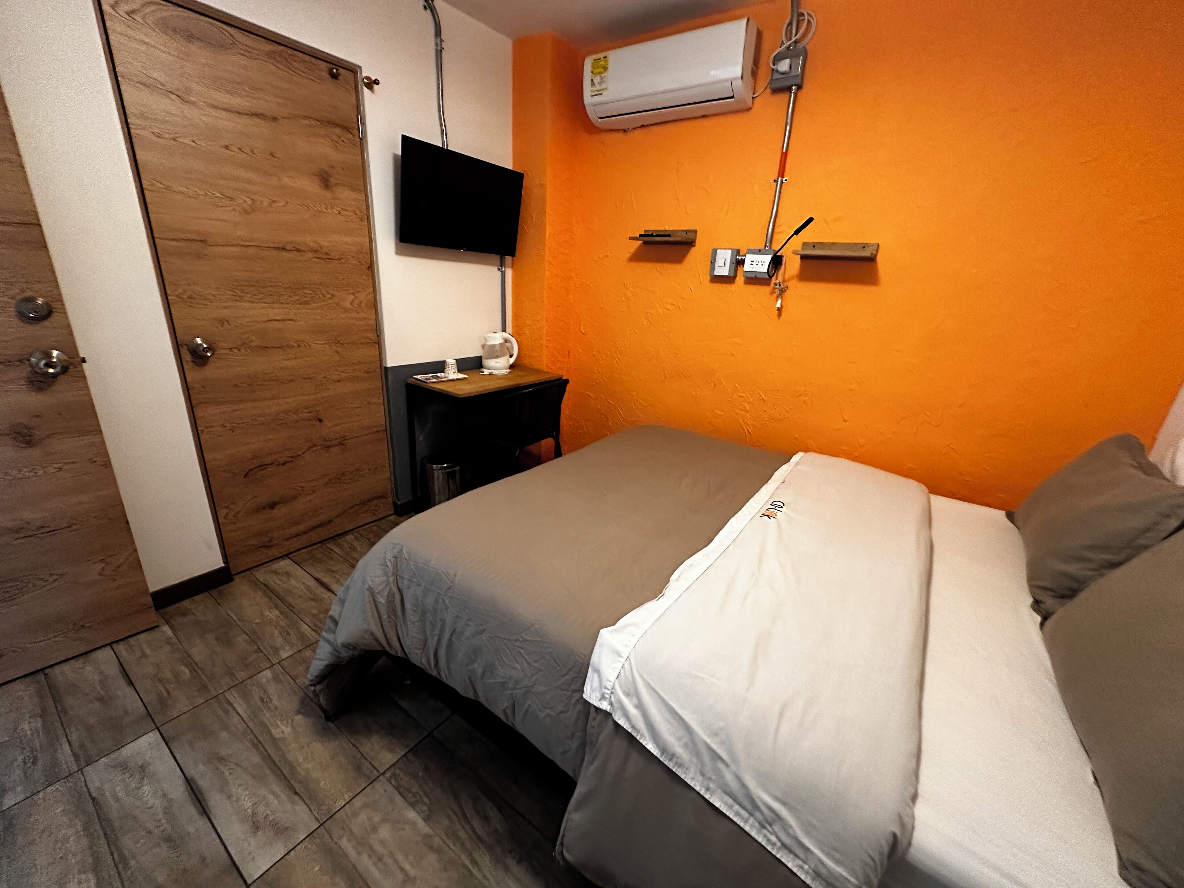 A bedroom with a wooden door, a flat-screen TV mounted on the wall, an air conditioning unit, a bedside table with a lamp, and a bed with a gray and white duvet.