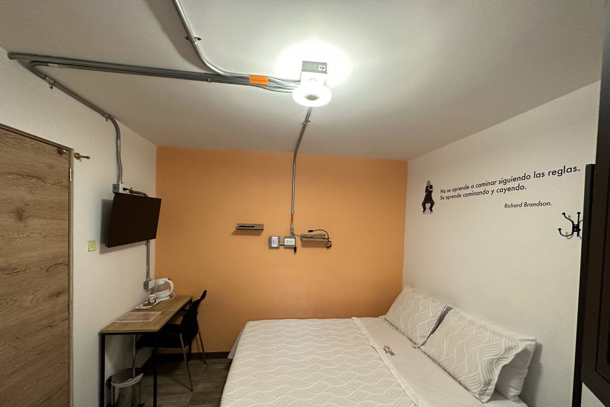 Ein kleiner, gut beleuchteter Raum mit einem Bett, einem Schreibtisch und einem an der Wand befestigten Fernseher.