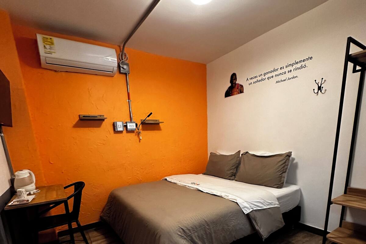 Ein kleines Hotelzimmer mit einem Bett, einem Schreibtisch mit Stuhl, einem kleinen Tisch und einem an der Wand befestigten Regal.