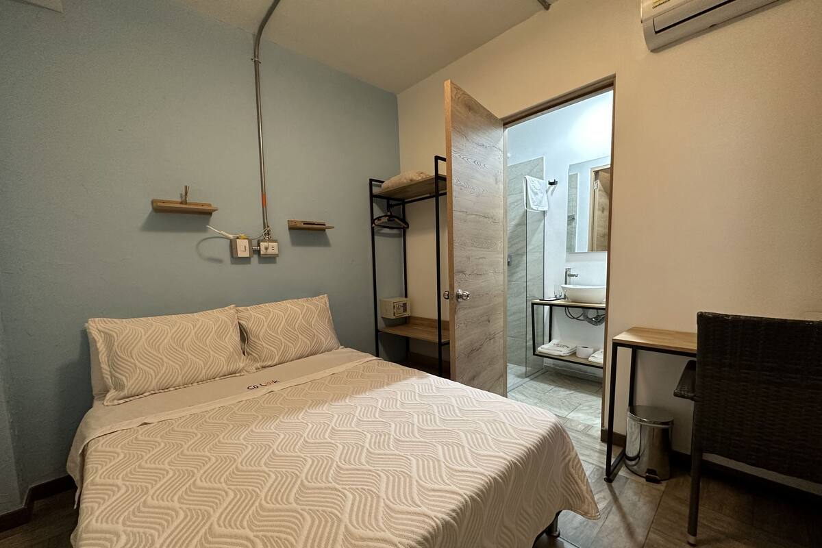 Ein Schlafzimmer mit Bett, Nachttischen, Schreibtisch und angrenzendem Badezimmer, das durch eine geöffnete Tür sichtbar ist.