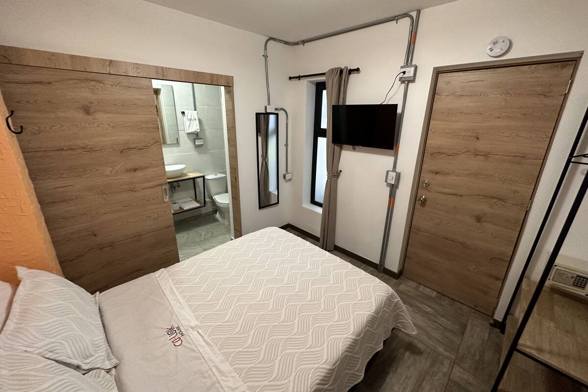 Ein Hotelzimmer mit Bett, Wandfernseher, Holztür und einem durchgegangene Tür erkennbaren Badezimmer.