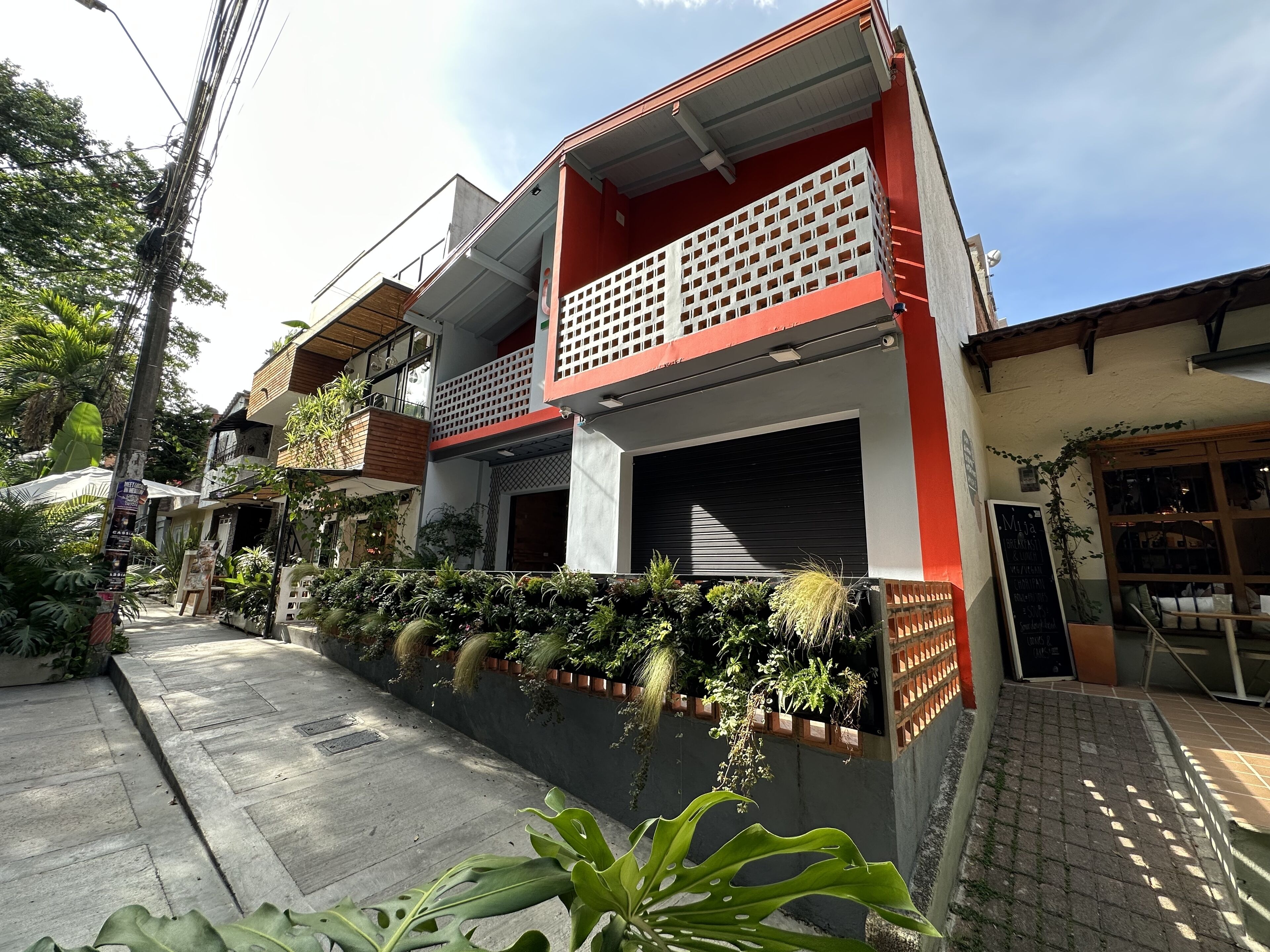 A modern two-story building with a red and white facade, a balcony with a lattice design, and a variety of potted plants along the front.