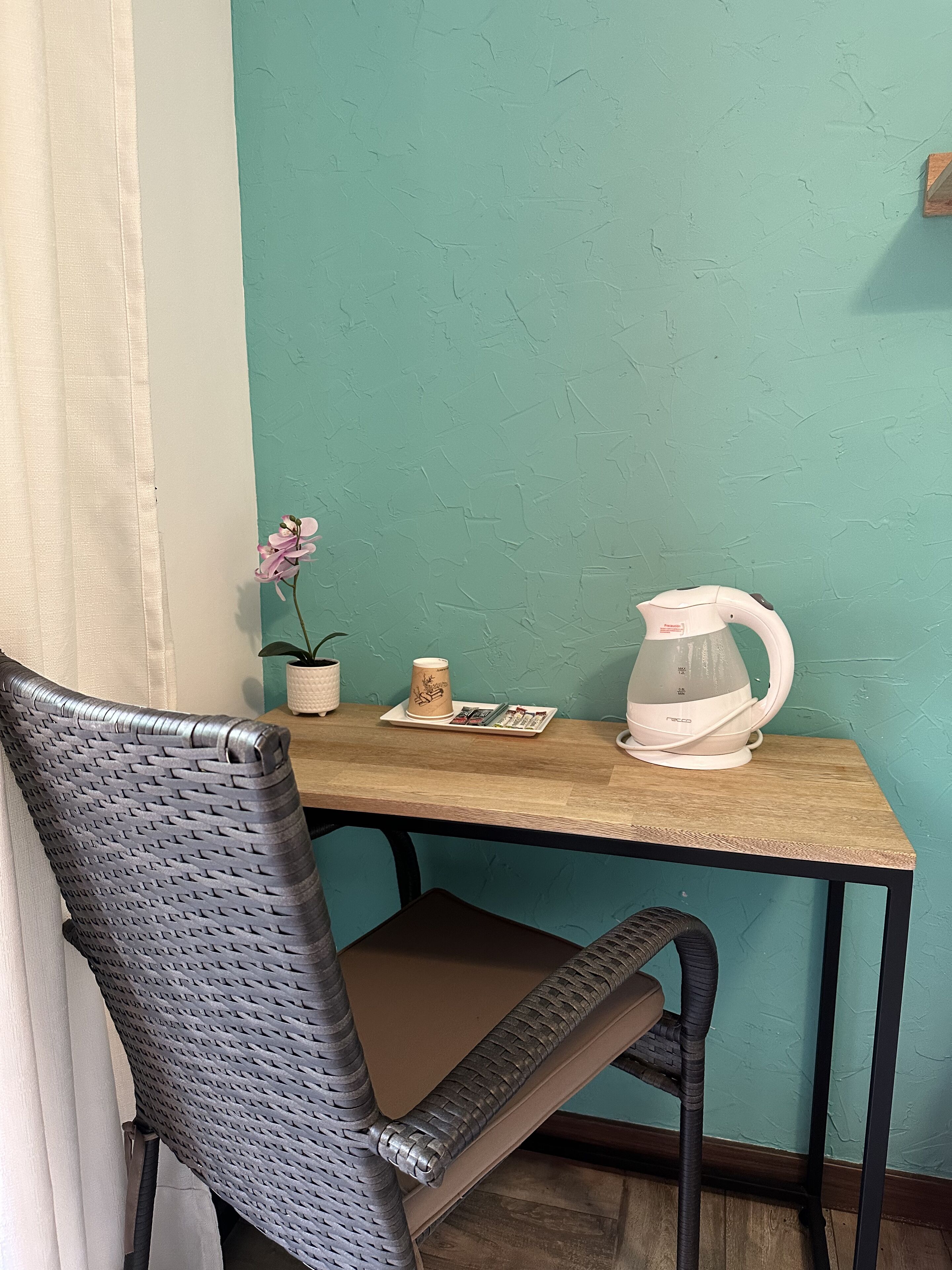 A corner of a room with a teal wall, a wooden table, a wicker chair, a small potted plant, and a white kettle.