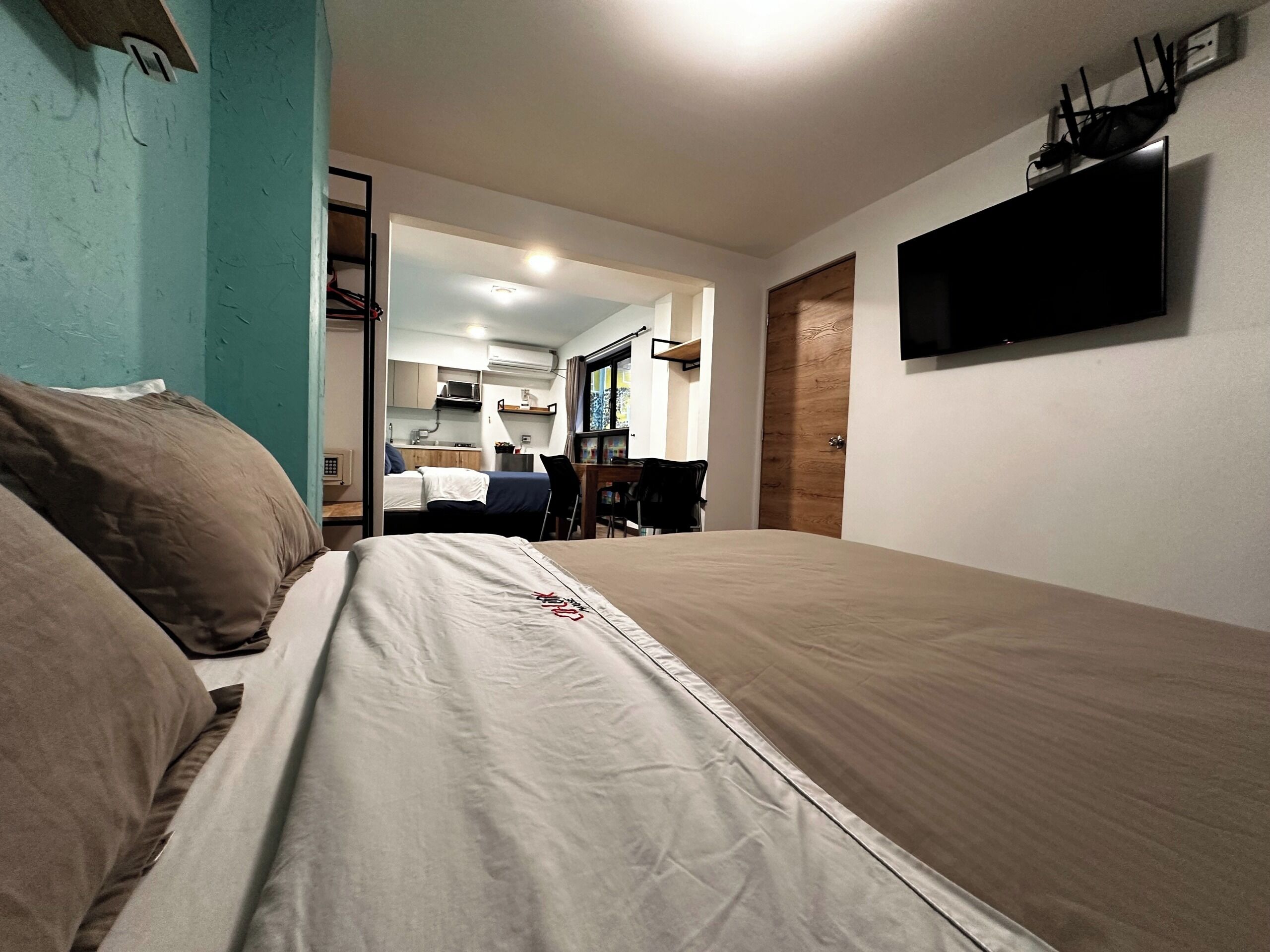 Un dormitorio con cama, televisor montado en la pared y una kitchenette al fondo.