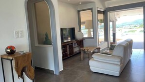 Ein modernes Wohnzimmer mit einem weißen Sofa, einem Holztisch, einem Fernseher auf einem Holzständer und einem großen Fenster mit Blick ins Freie.