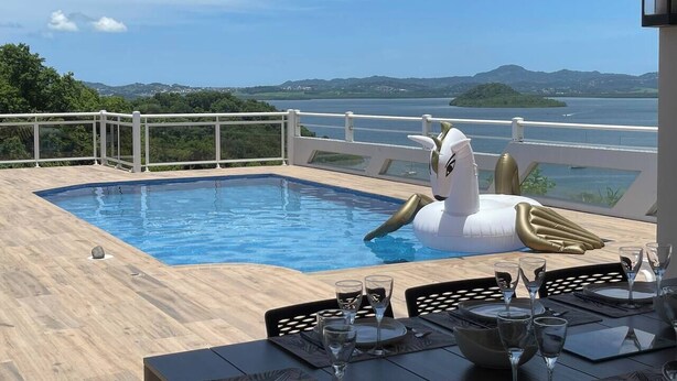 Ein Poolbereich mit Blick auf das Meer und die Hügel, ein weißer aufblasbarer Poolreifen und eine Esszeile mit Gläsern und Besteck.