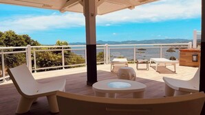Ein überdachter Außenbereich mit weißer Möbel, einem runden Tisch und Blick auf das Meer und hügelige Landschaft.