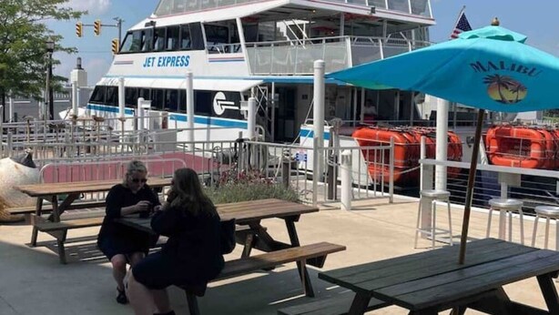 En Jet Express færge ligger til kaj ved en bro, med udendørs siddepladser og en parasol med Malibu-tema.