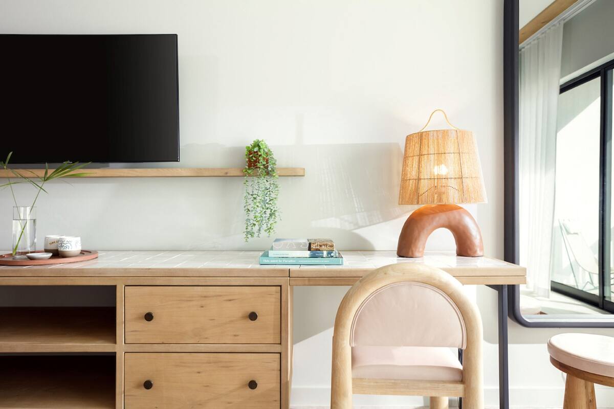 A modern workspace with a large flat-screen TV, a wooden desk, a chair, a lamp, and a window with a view.