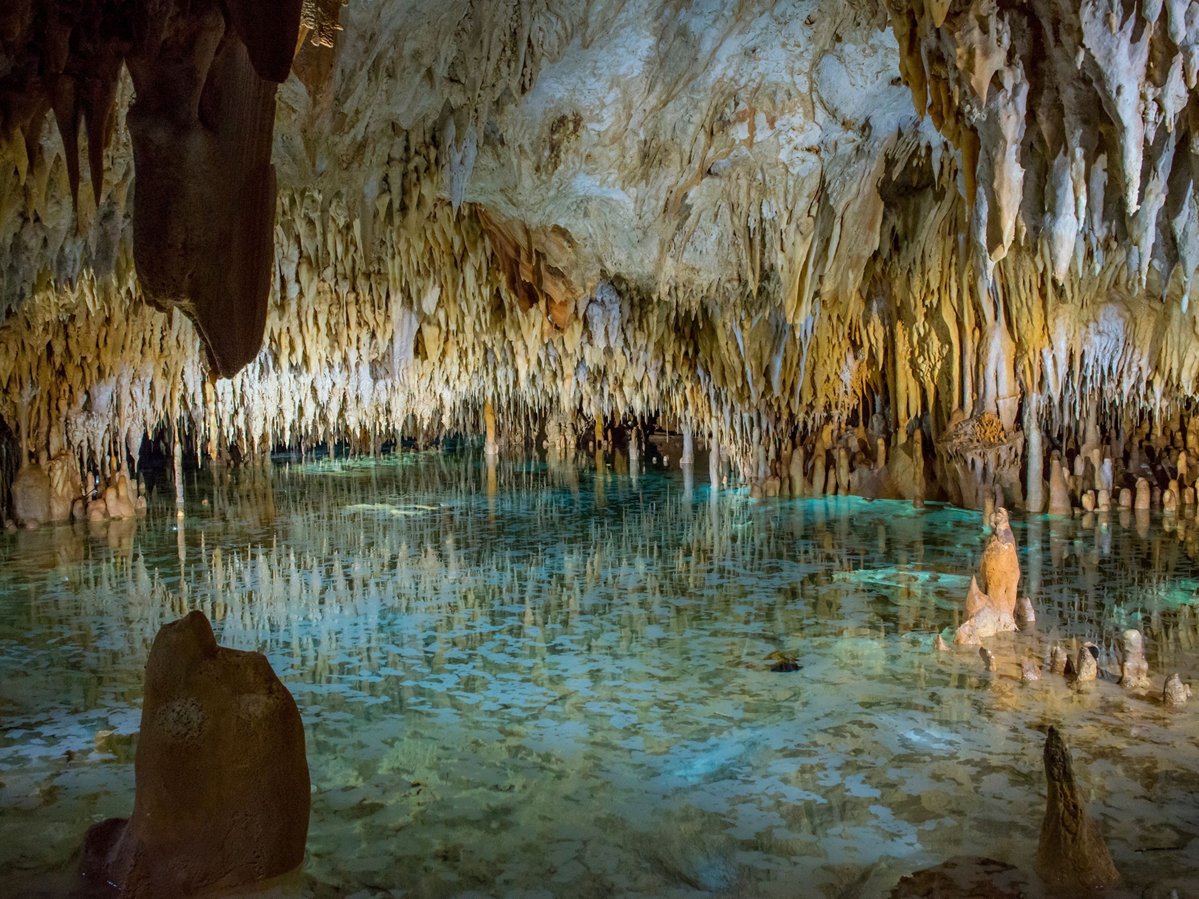 A cave with stalactites and stalagmites, clear water, and rock formations.