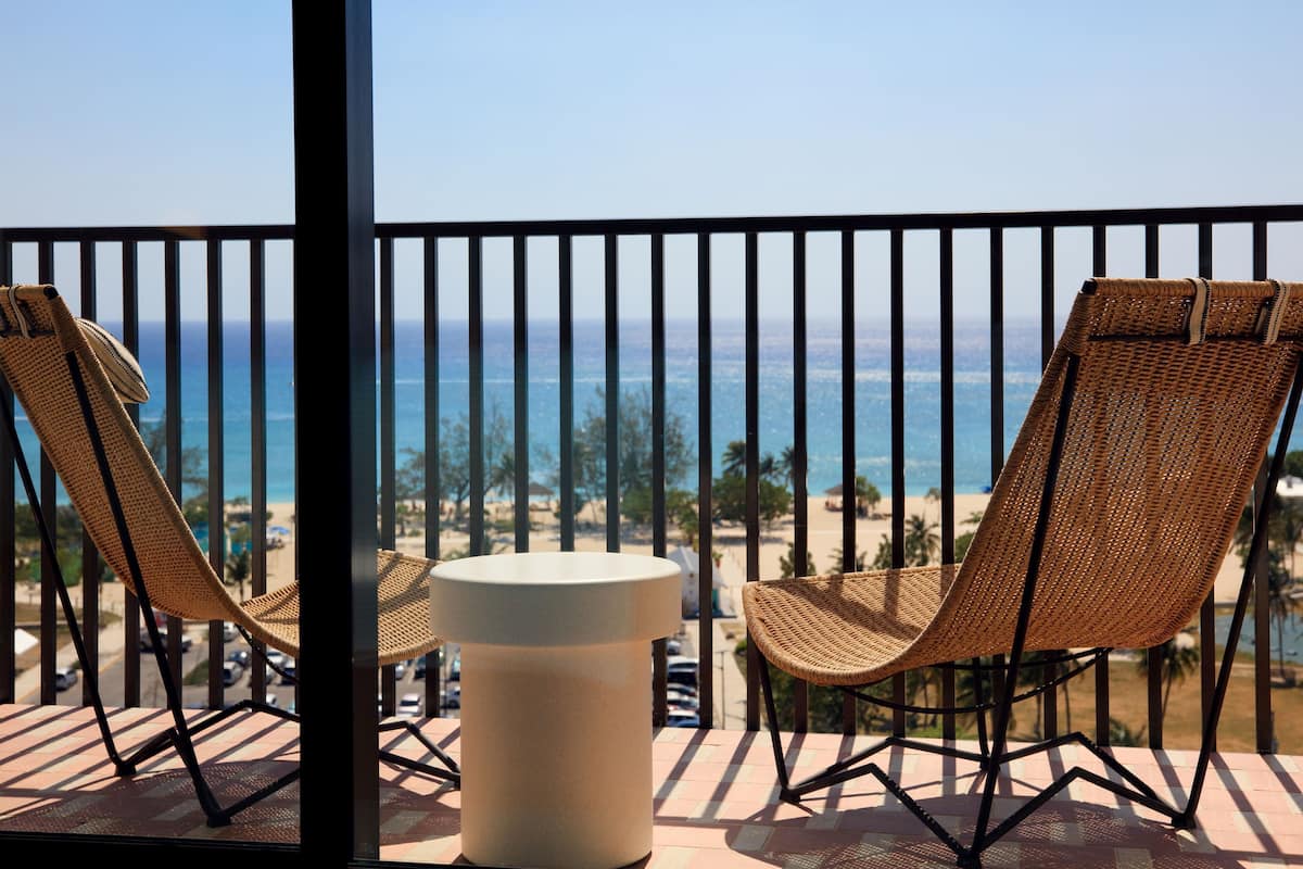 A balcony with two wicker chairs and a round table, overlooking a beach and ocean.
