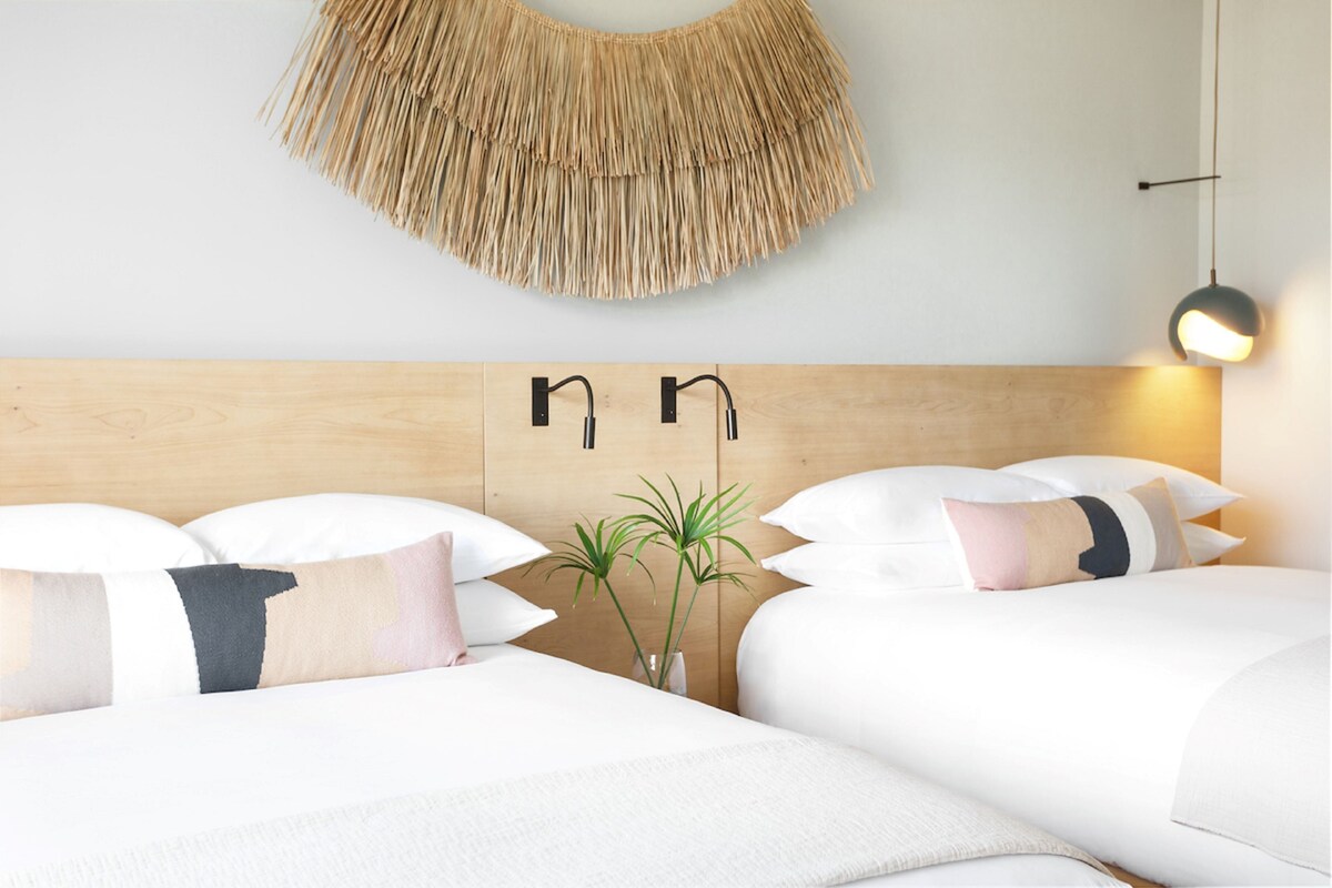 Habitación de hotel con cabecera de madera, dos camas con ropa blanca, una planta pequeña y un aplique de pared.